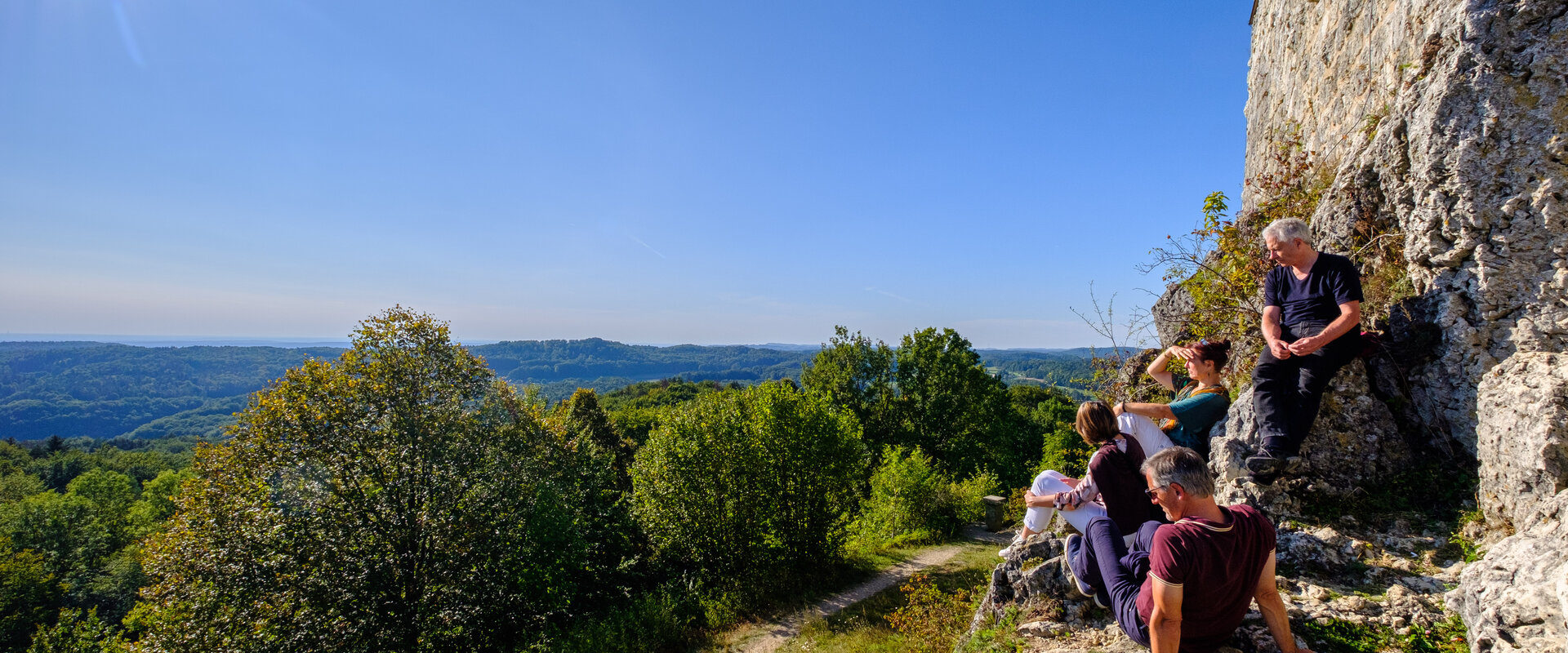 Wanderer sitzen auf den Felsen vor der Burg Hohenstein und schauen bei blauem Himmel und Sonnenschein in die hügelige und waldige Landschaft